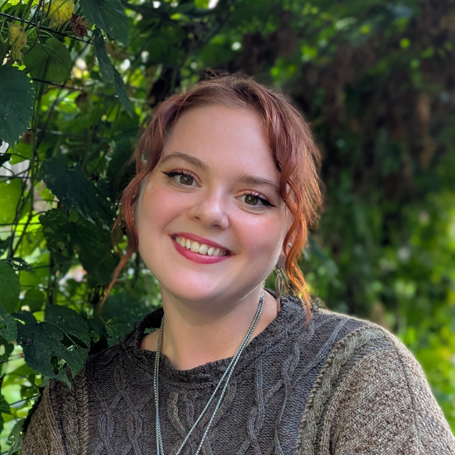 A photograph of Victoria standing in front of cascading green leaves.